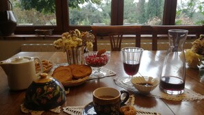 Una mesa dispuesta con un calentador de agua, taza de té, azucarero, pan y una copa de vino tinto, con vistas a árboles a través de la ventana.
