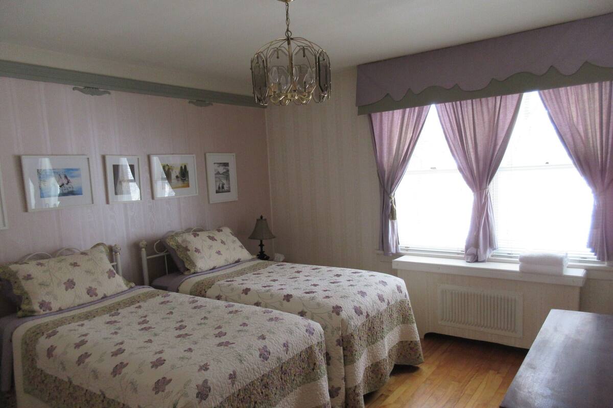 Ein Schlafzimmer mit zwei Betten, einem Heizkörper, einem Fenster mit Vorhängen und einer Kristallleuchtre.