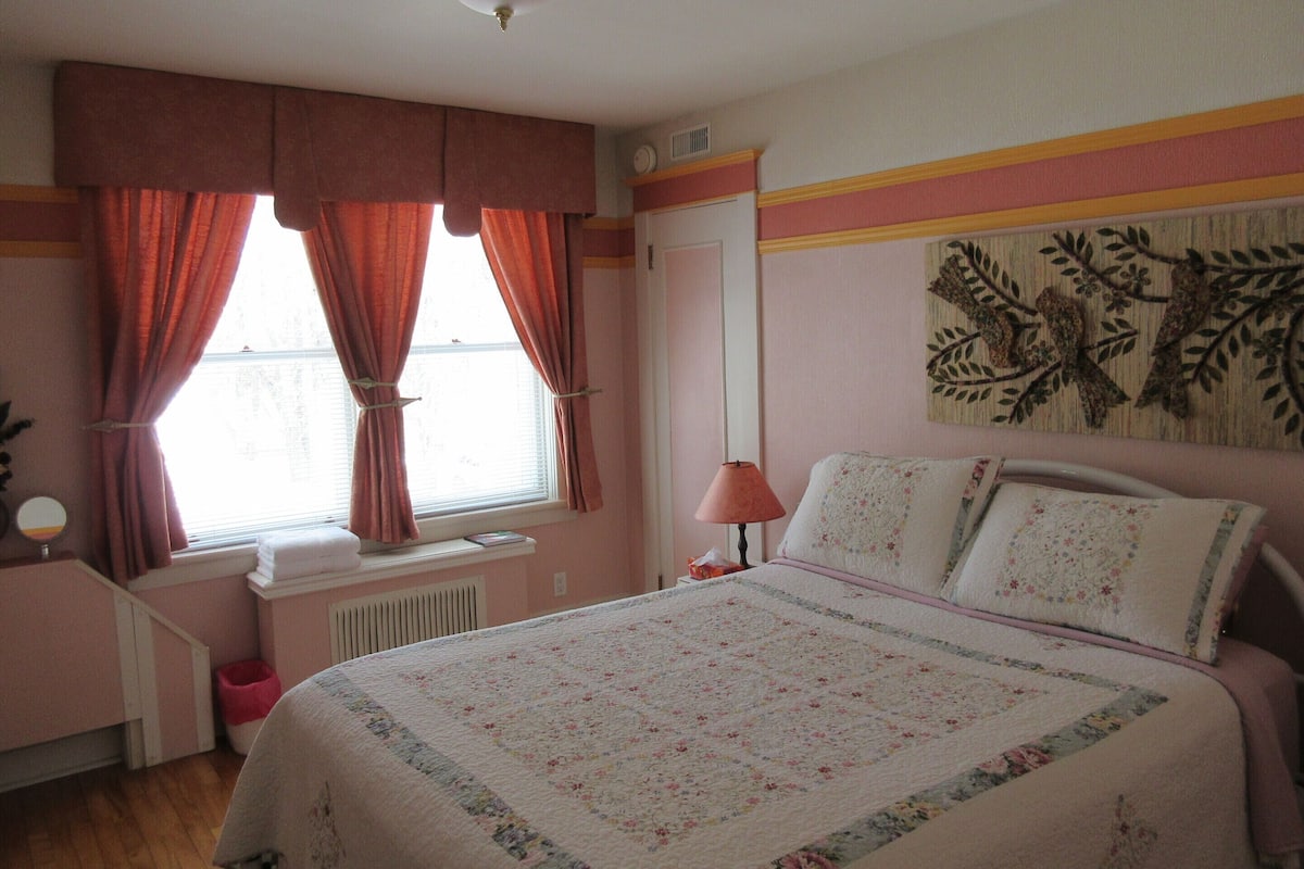 Ein Schlafzimmer mit Bett, einem Fenster mit Vorhängen, einem Heizkörper, einer Wanduhr und einem Wanddekor.