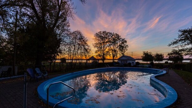 Una piscina con un paseo de ladrillos alrededor, árboles y un pabellón al fondo.