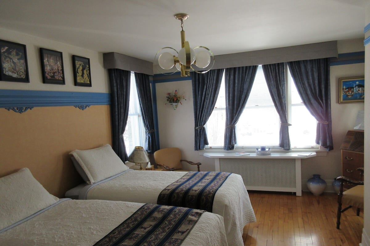 Ein Hotelzimmer mit zwei Betten, einem Schreibtisch, einem Stuhl, einem Heizkörper und einem fenster mit Vorhängen.