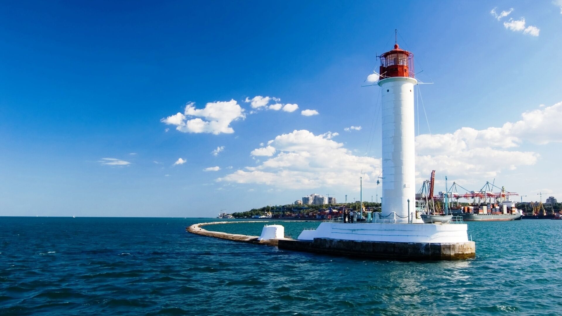 A lighthouse on a pier with a clear view of the sea and a distant cityscape.