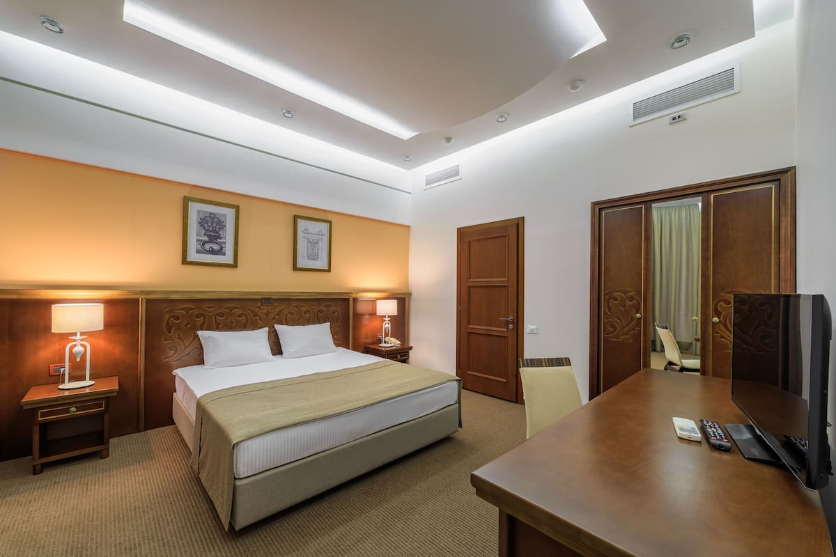 A hotel room with a bed, bedside lamps, a desk with a TV, and a door.