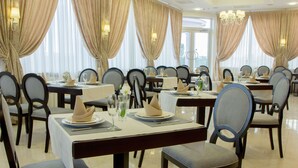 A formal dining area with multiple tables set for service, featuring chairs with cushioned seats and backrests, and a chandelier hanging from the ceiling.