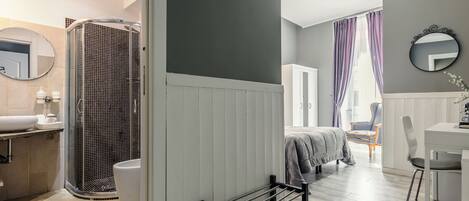 Un baño moderno con ducha, inodoro y lavamanos. En la habitación contigua hay una cama y un escritorio.