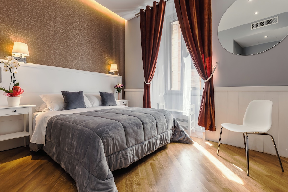 A modern bedroom with a large bed, a bedside table with a lamp, a chair, and a window with curtains.