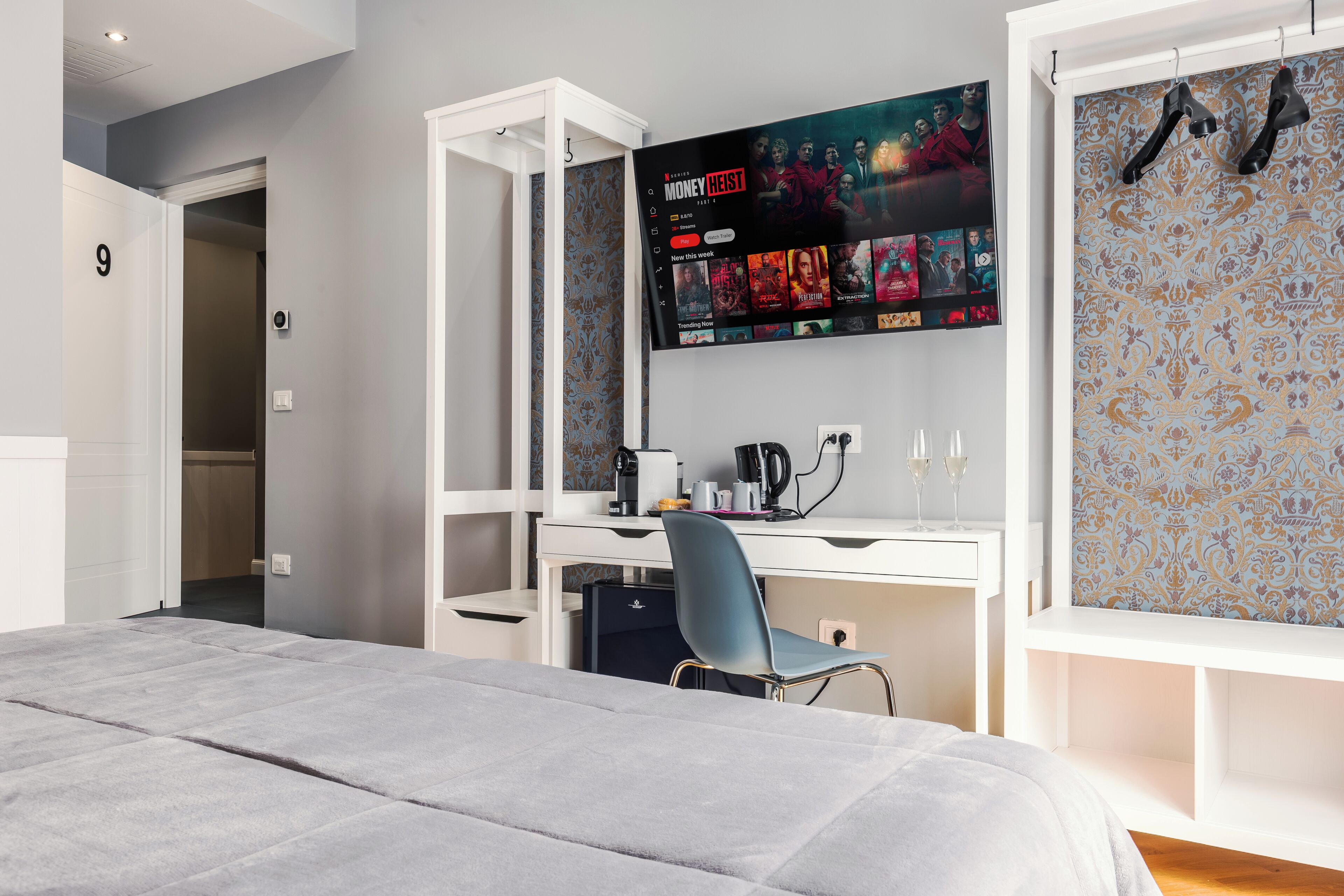 Quarto de hotel moderno com uma cama grande, uma escrivaninha com computador, uma TV montada sobre a escrivaninha exibindo um filme e um papel de parede estampado.