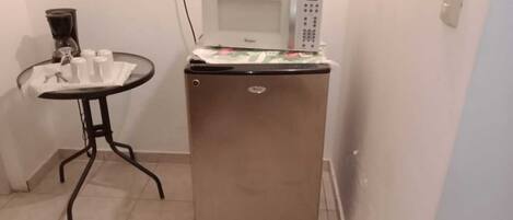 A compact kitchenette with a microwave on top of a refrigerator, a small table with cups and a pitcher, and a tiled floor.