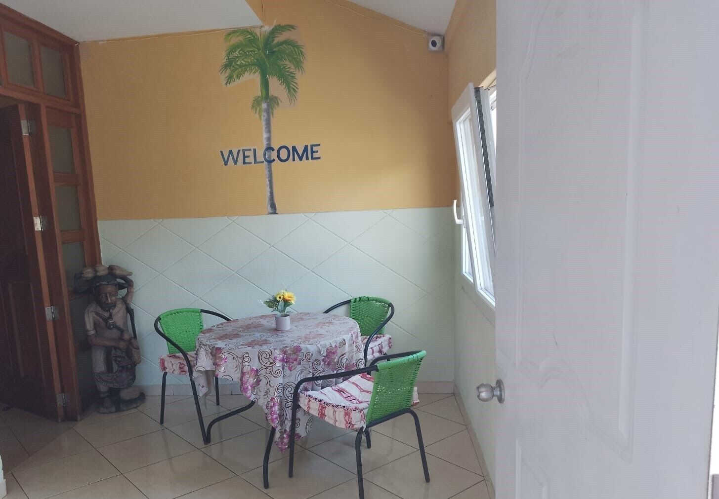 A room with a 'WELCOME' sign, a statue, a table with chairs, and a window.