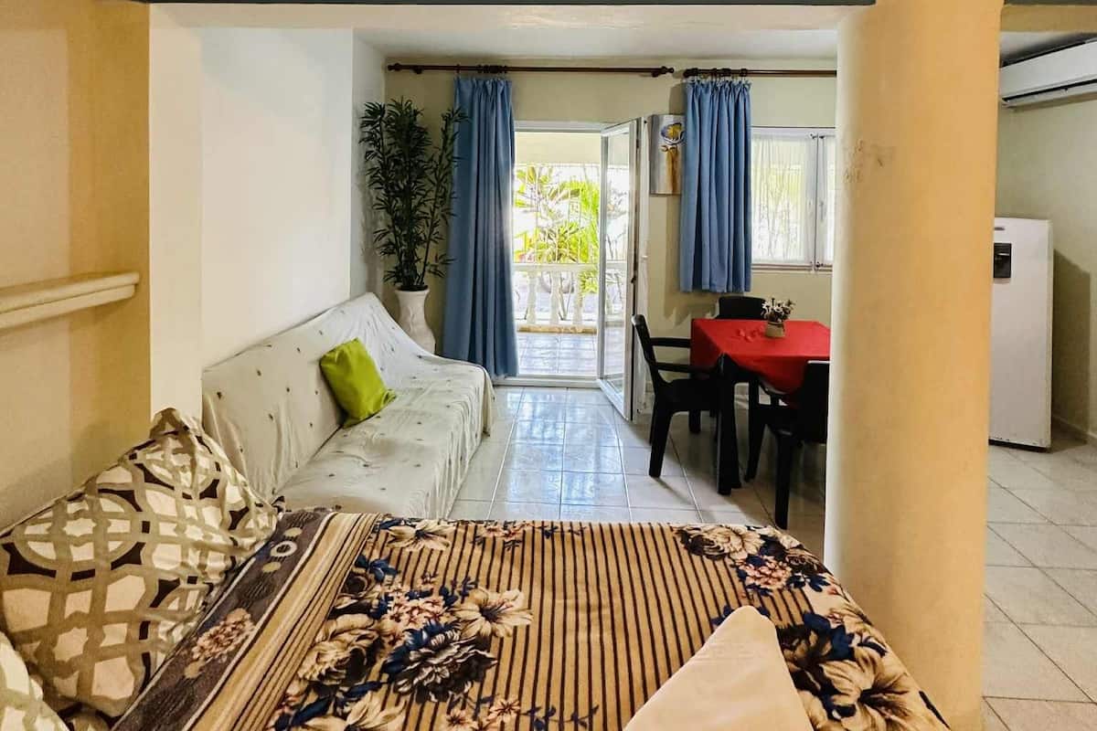 A compact living space with a sofa, a dining table with chairs, a small kitchen area, and a view through a glass door.