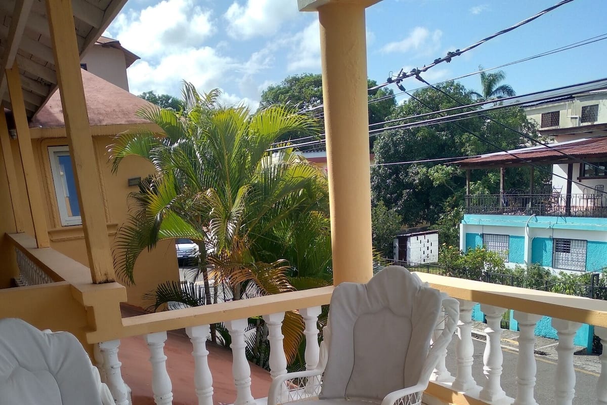 A balcony with white wicker furniture, including chairs and a table, and a view of the surrounding area with buildings and trees.