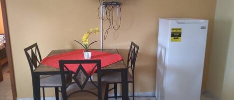 A dining area with a table and chairs, a television mounted on the wall, and a refrigerator.