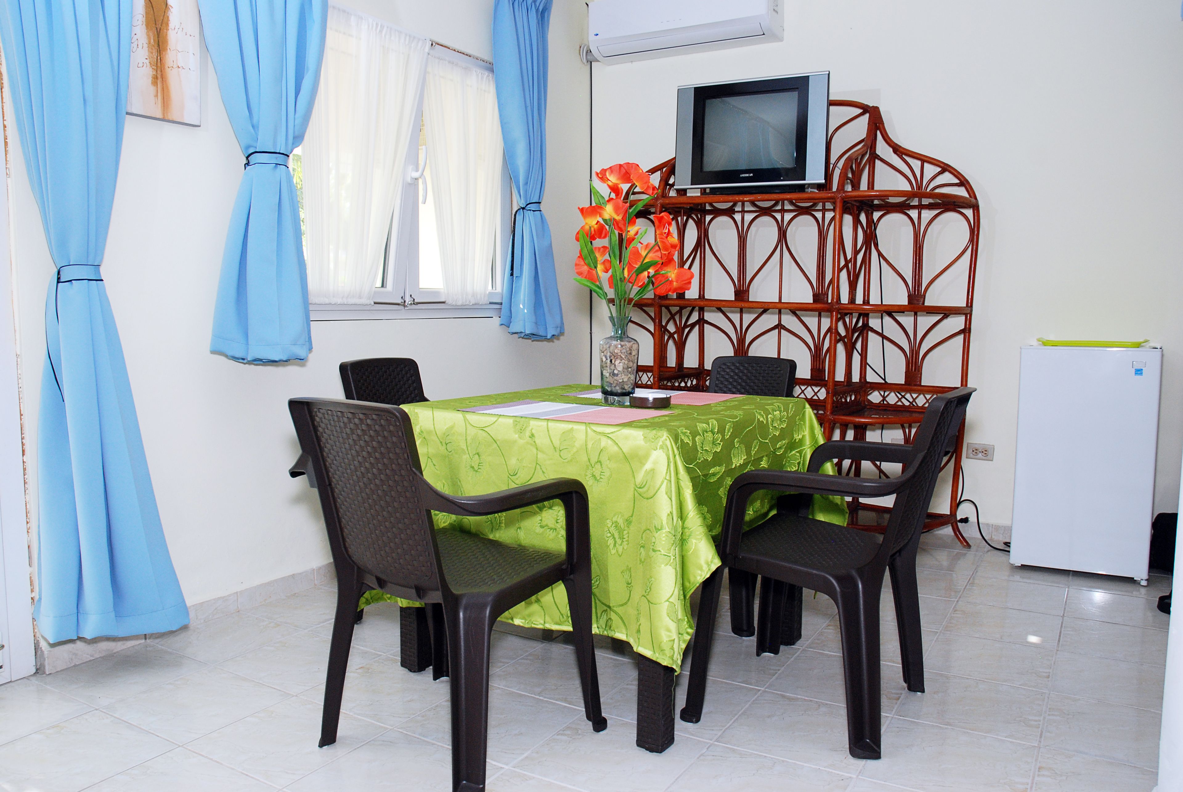 Comedor con una mesa dispuesta para seis personas, rodeada de sillas, un televisor sobre un mueble bajo y un refrigerador.