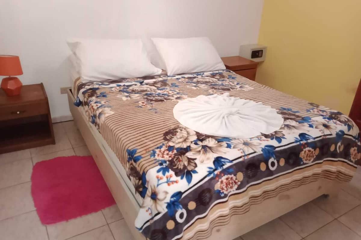 A neatly made bed with a floral bedspread and a white towel, a wooden nightstand with a lamp, and a red rug on the floor.