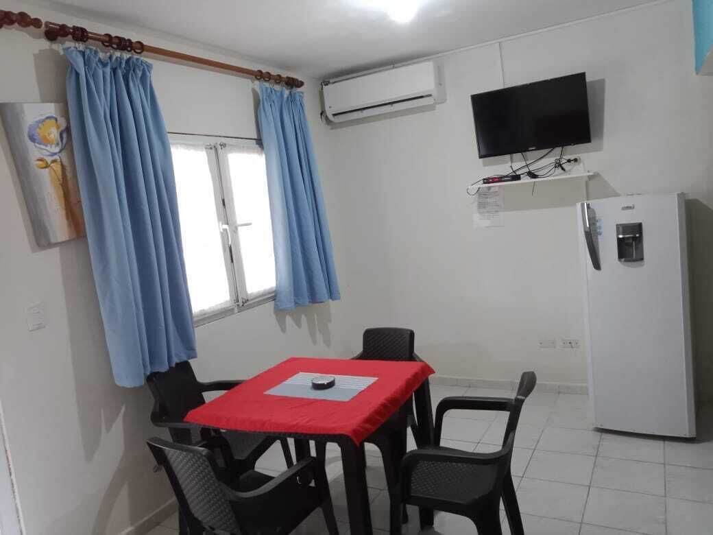 Quarto pequeno com uma mesa, cadeiras, televisão e frigorífico.