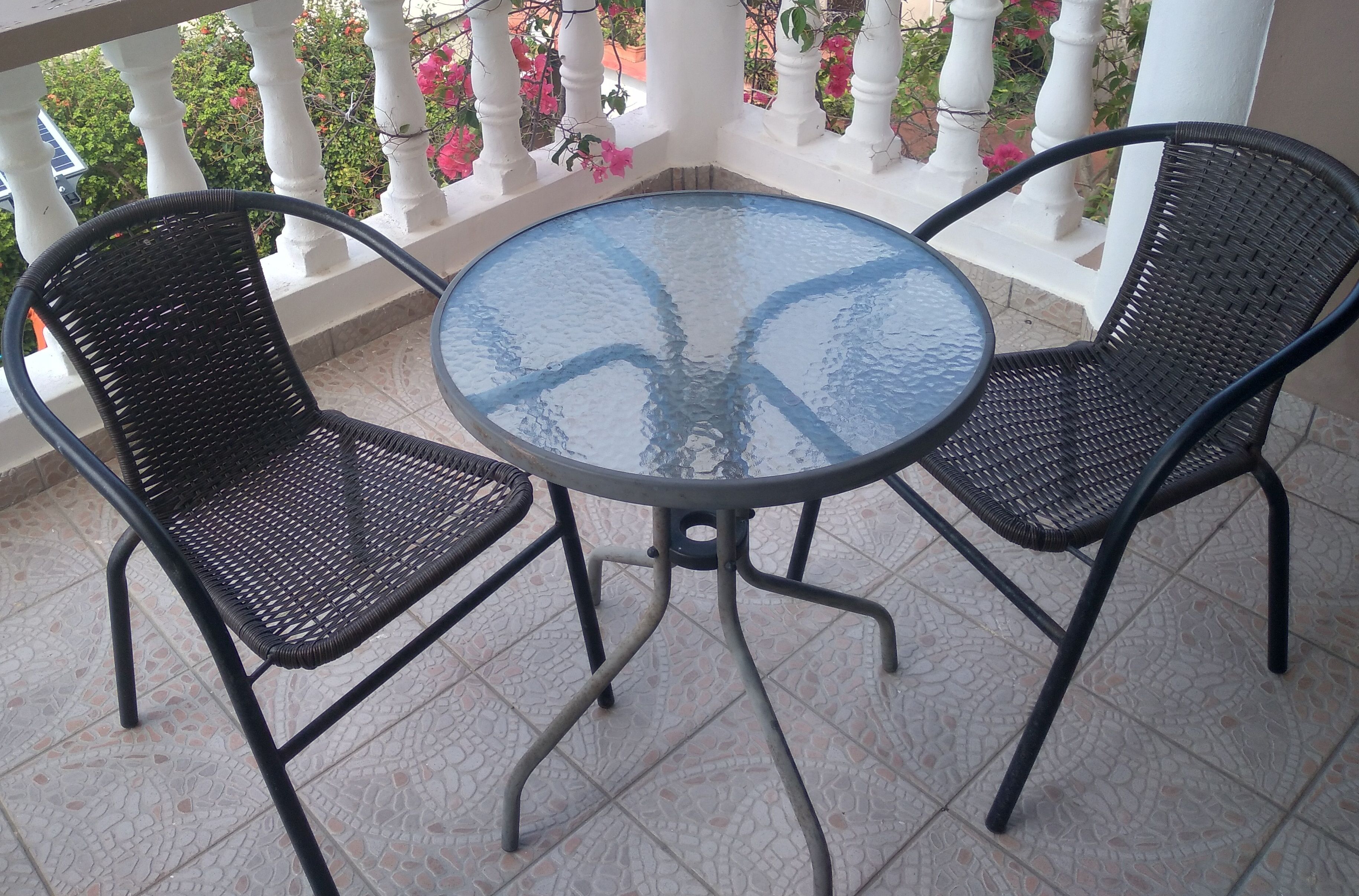 A round glass table with two black wicker chairs on a tiled balcony with white balustrades and pink flowers.