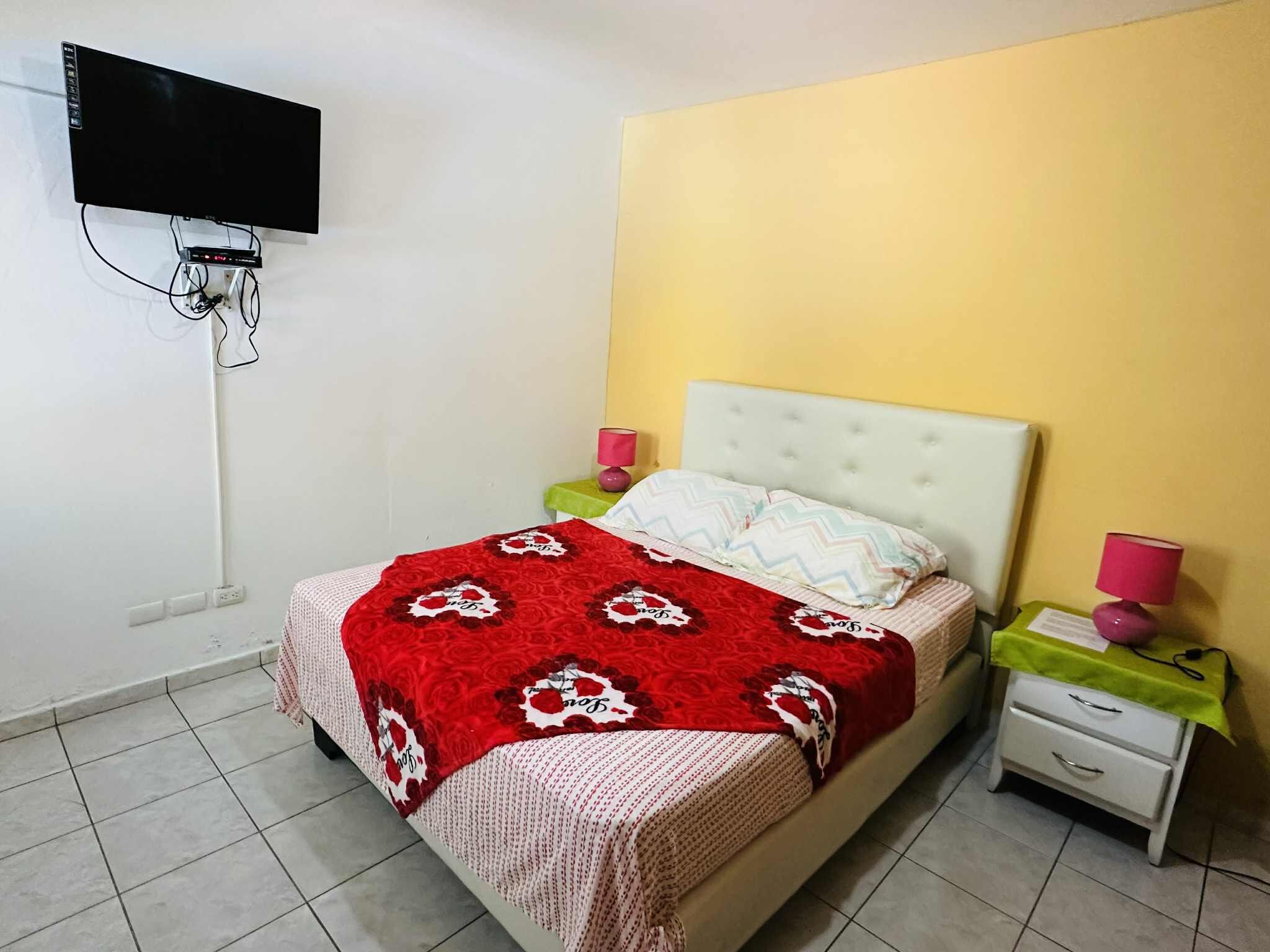 Un dormitorio con cama, mesitas de noche, televisor montado en la pared y una pared amarilla.