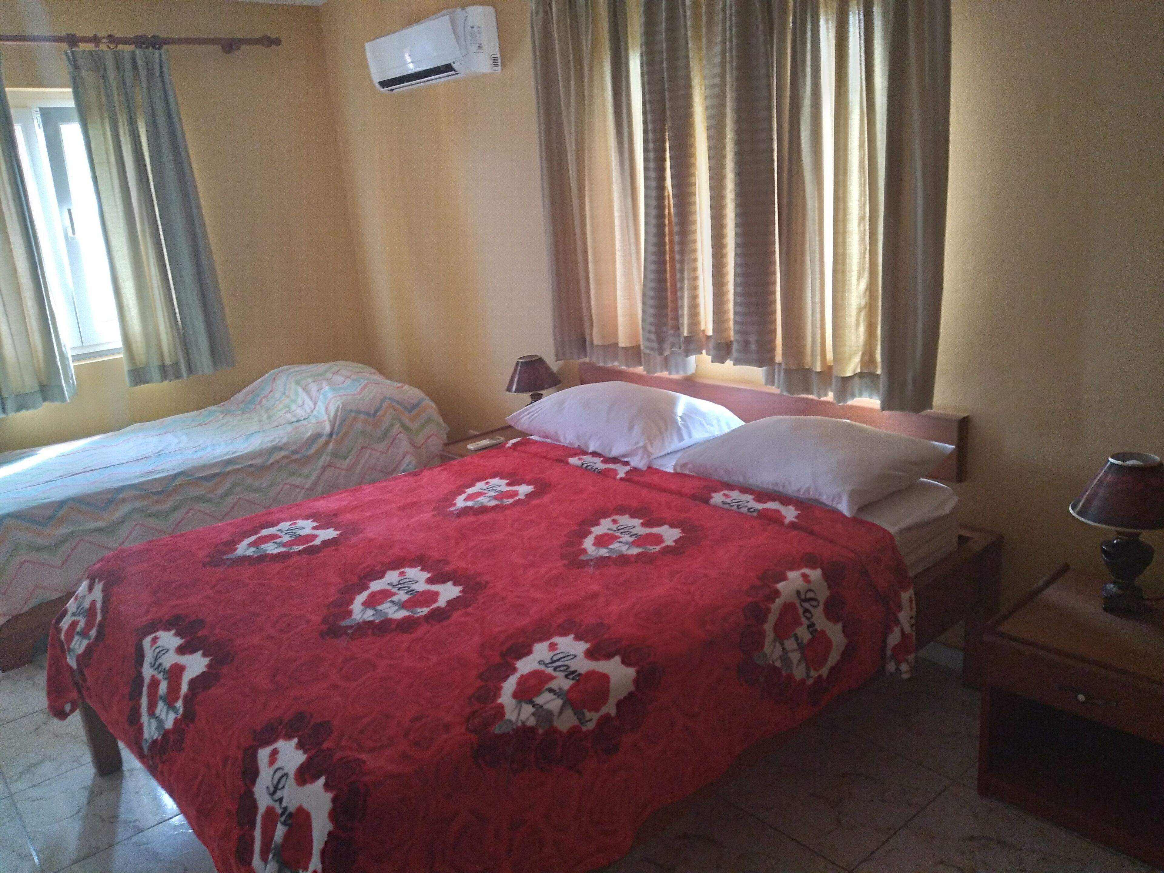 A hotel room with two beds, one with a red floral bedspread and the other with a striped bedspread. There is a window with curtains, an air conditioning unit on the wall, and a nightstand with a lamp.