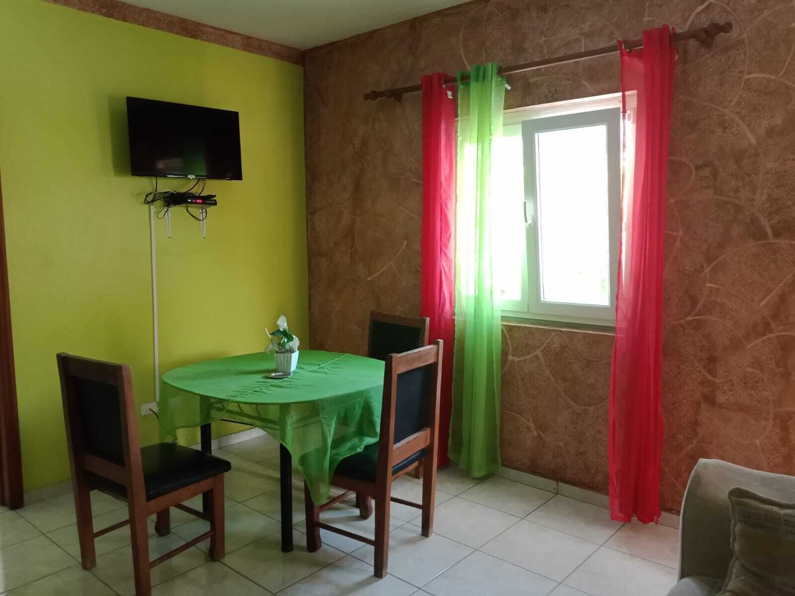 A dining area with a green table and chairs, a flat-screen TV mounted on the wall, and a window with red and green curtains.