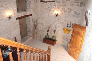 Interior rústico de piedra con una escalera de madera, una pequeña mesa de madera con jarrones, y una puerta que dice 'LA POSADA'.