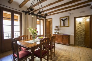 Un comedor con una mesa de madera, sillas y una araña. Hay ventanas con persianas, un jarrón con flores y una pared de piedra.