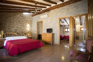 Un dormitorio con pared de piedra, vigas de madera, cama con ropa de cama roja, cómoda de madera, televisor y espejo.