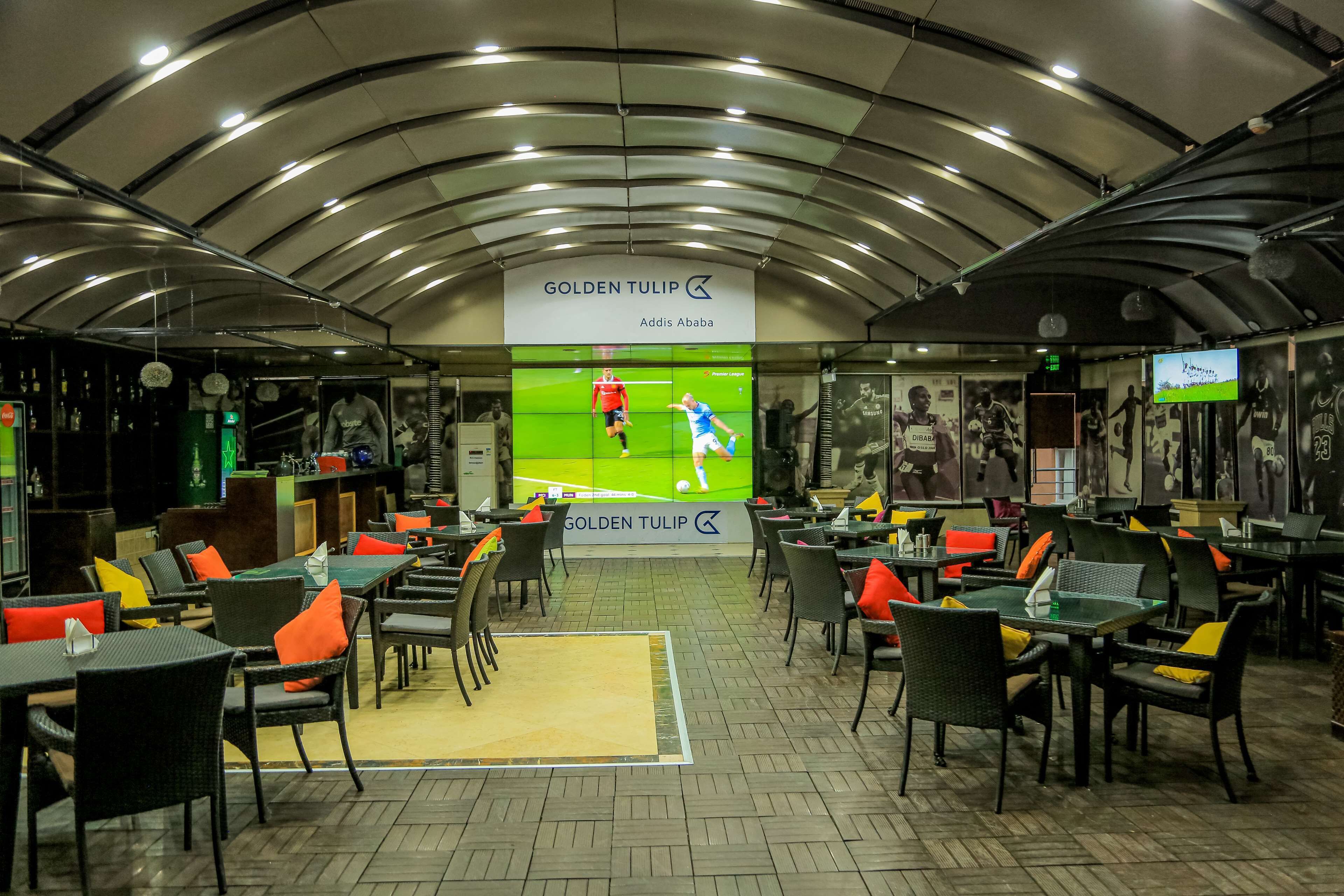 A restaurant with a sports-themed decor, featuring a large screen displaying a soccer game. Tables and chairs are arranged for dining.