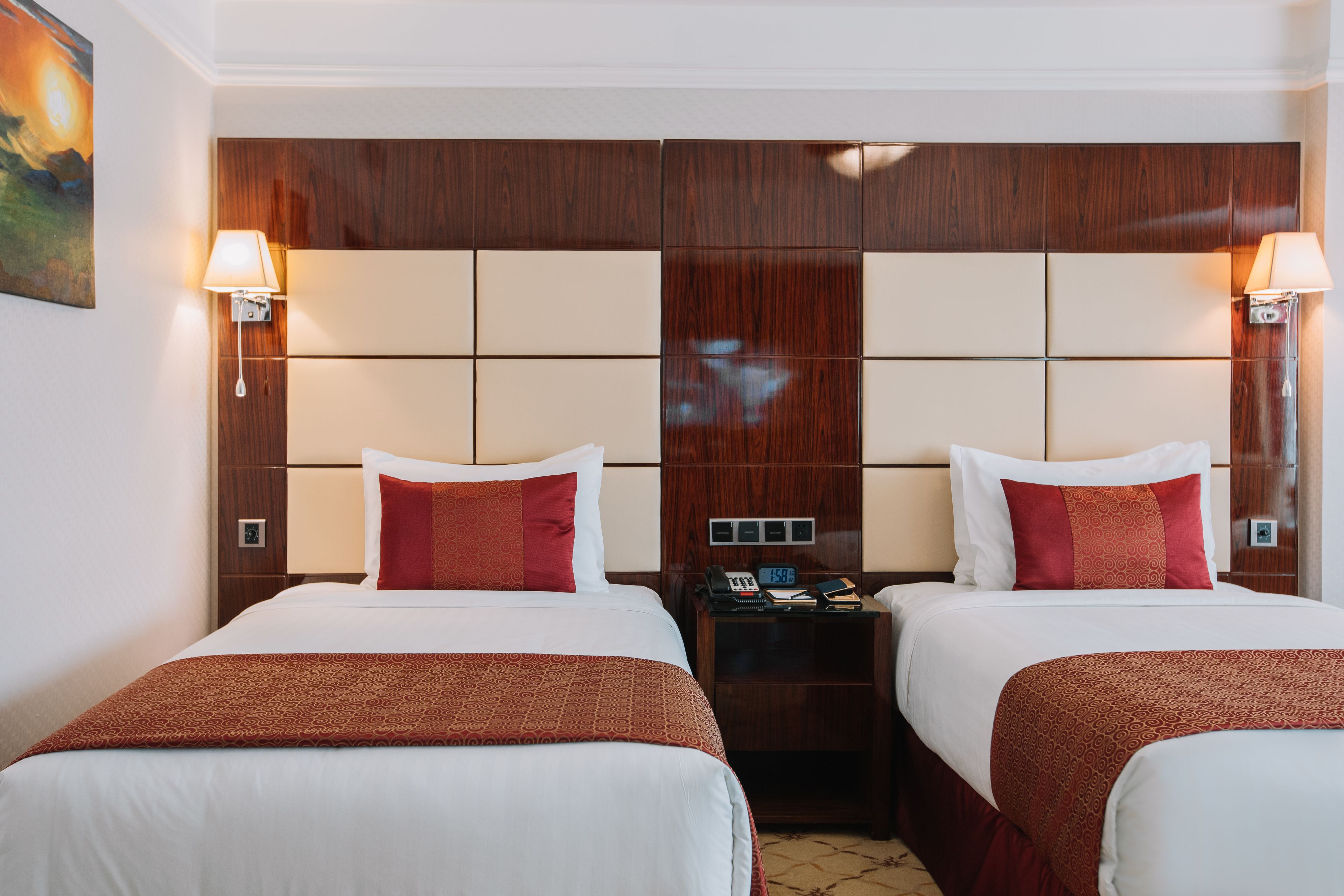 A hotel room with two beds, wooden headboards, bedside lamps, and a nightstand with a phone and clock.