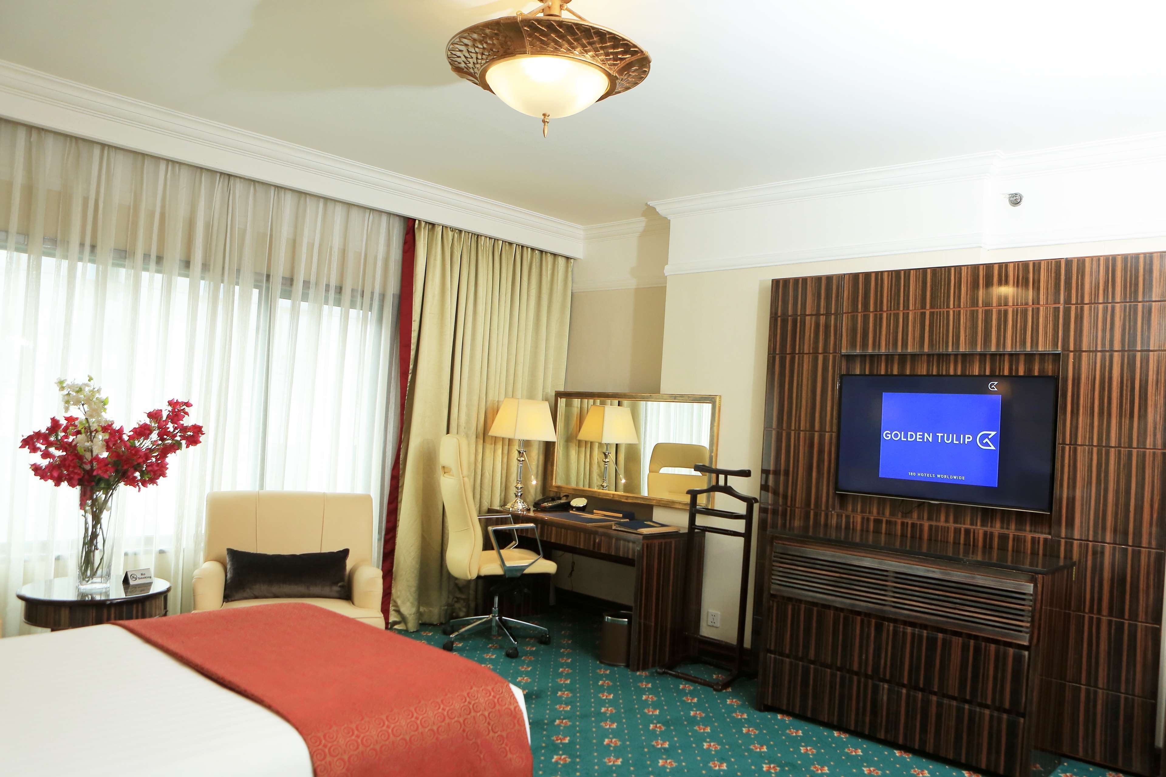 Ein Hotelzimmer mit Bett, Schreibtisch, Stuhl, Fernseher und einer Vase mit Blumen.