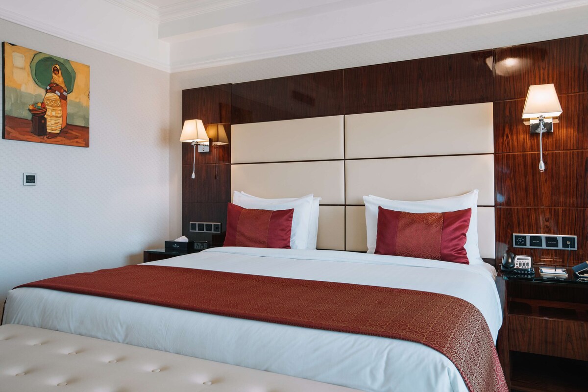 A hotel room with a large bed, two bedside lamps, a nightstand with a phone and a lamp, and a painting on the wall.