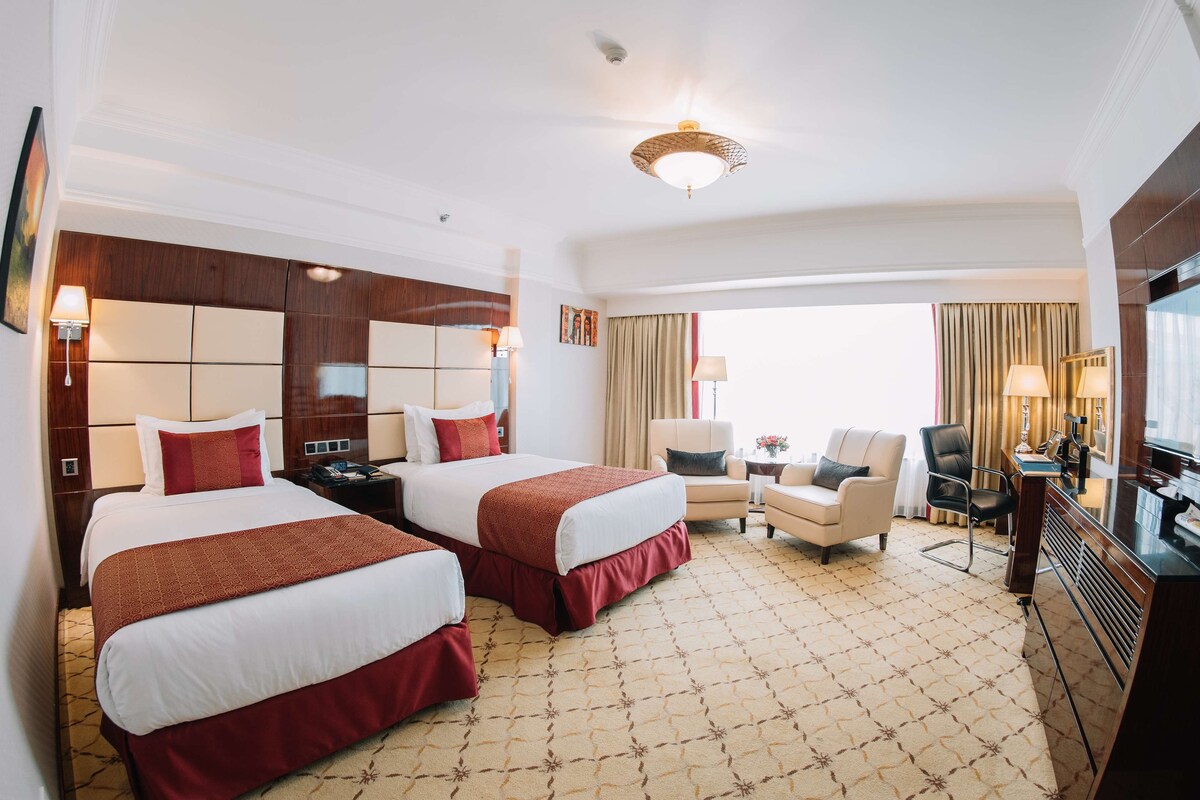 A hotel room with two beds, a desk, a chair, a TV, and a large window with curtains.