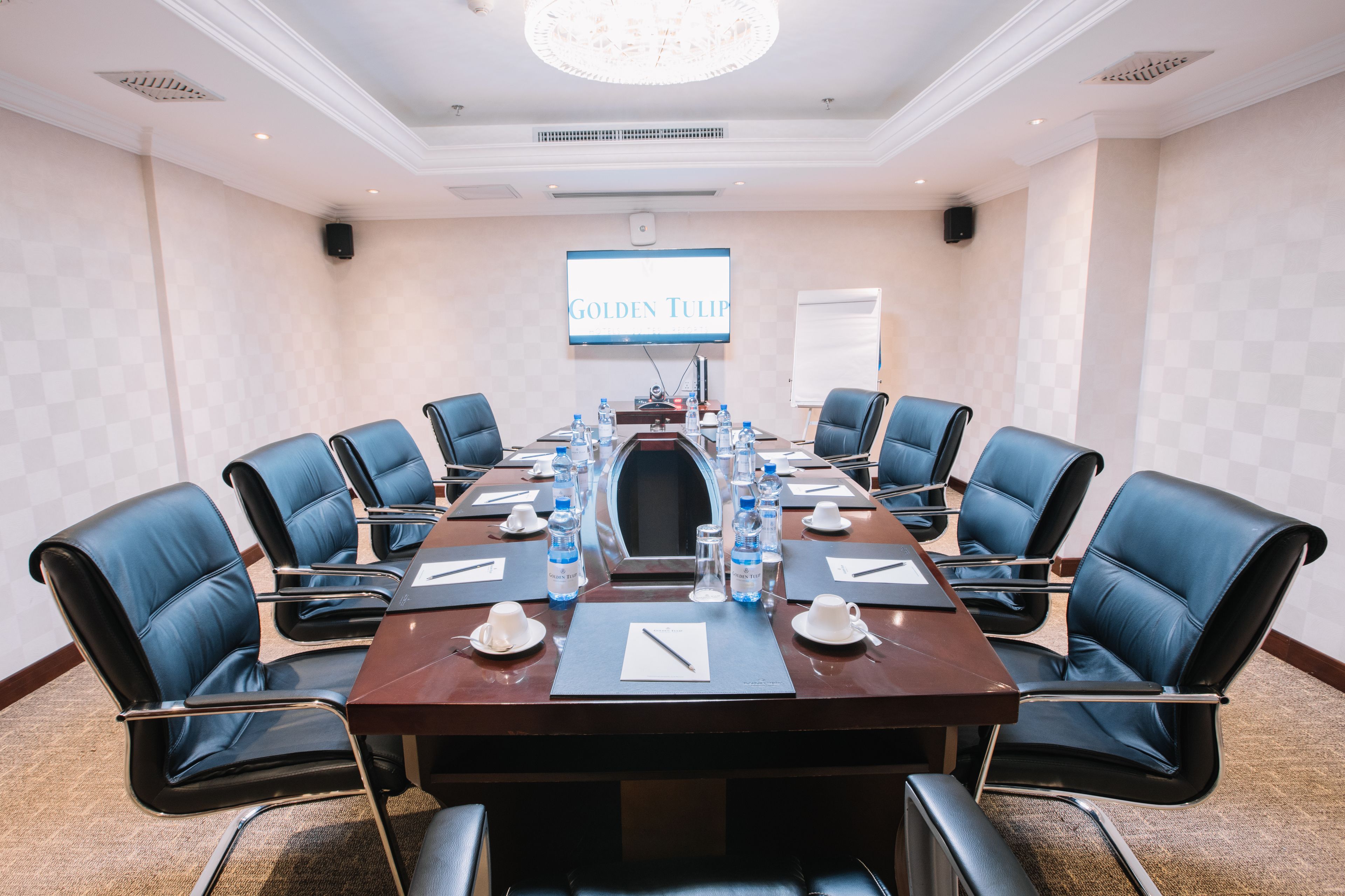 A conference room with a long table, blue leather chairs, water bottles, and a screen displaying "GOLDEN TULIP".