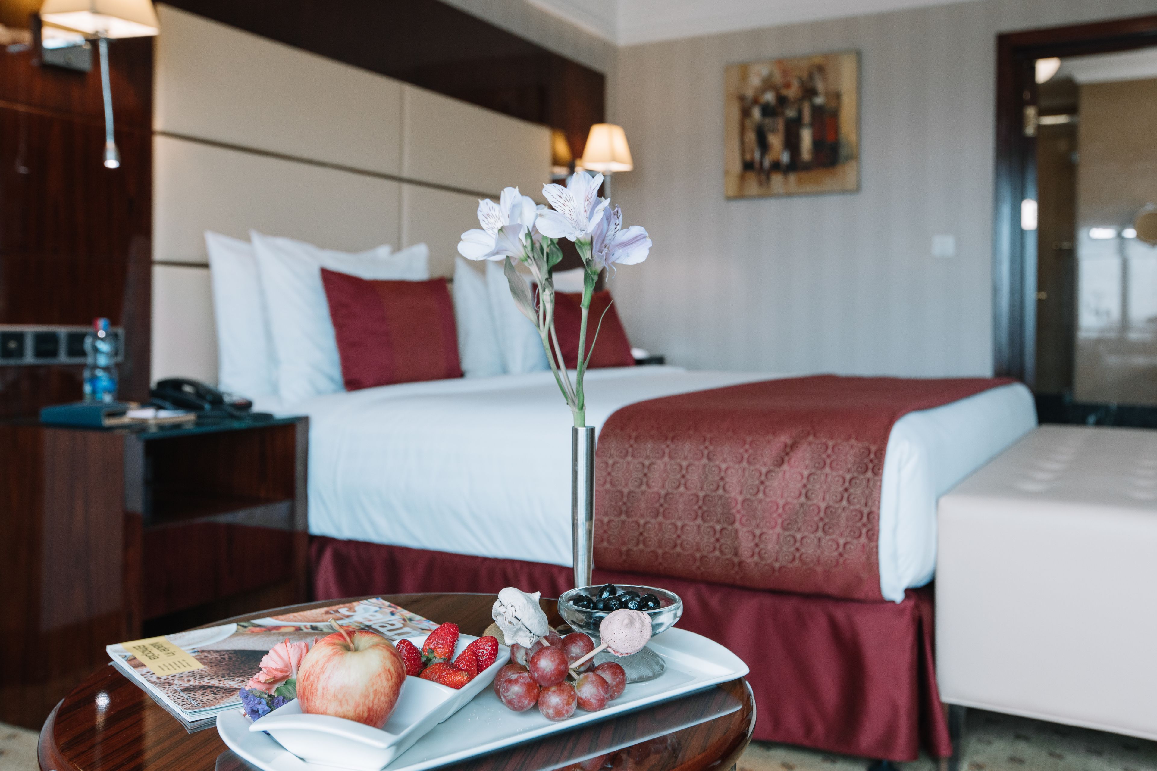 Ett hotellrum med en snyggt bäddad säng, ett nattduksbord med ett tidningsnummer, frukt och en vas med blommor.