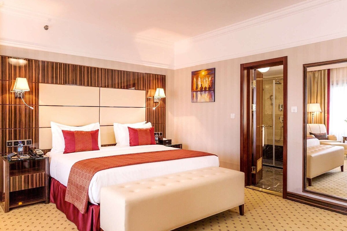 A hotel room with a large bed, a bench, a nightstand, a mirror, and a shower area.