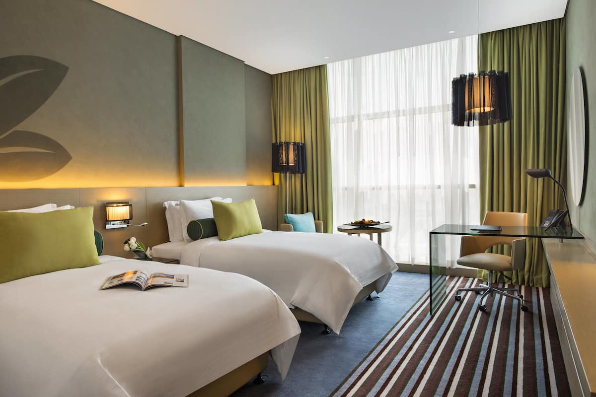A hotel room with two beds, a desk, a chair, a small table with fruit, and a large window with curtains.