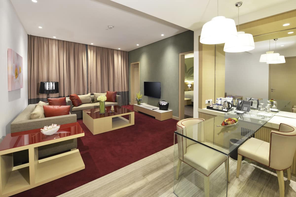 A modern living room with a sofa, coffee table, and a glass dining table with chairs. There is a television and a kitchen area with a coffee machine and a kettle.