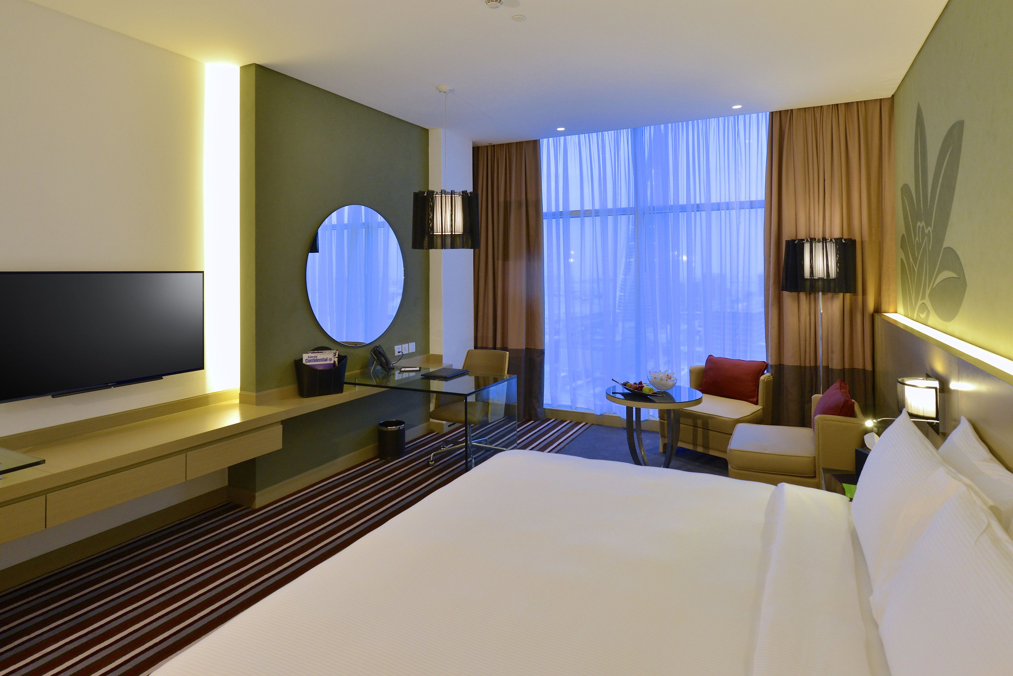 Habitación de hotel con una cama grande, televisor, escritorio, silla, mesita y ventana con cortinas.