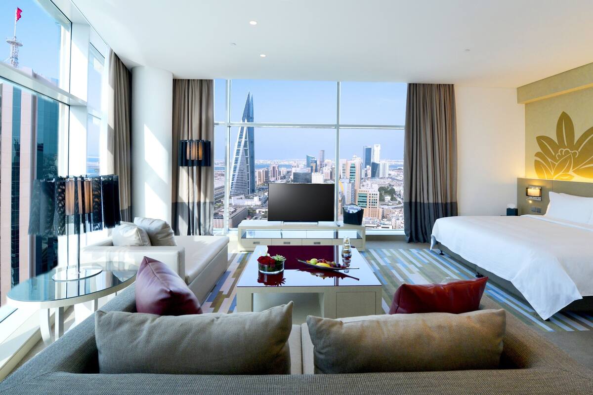 A hotel room with a large window, a bed, a sofa, a TV, and a small table with a fruit bowl.