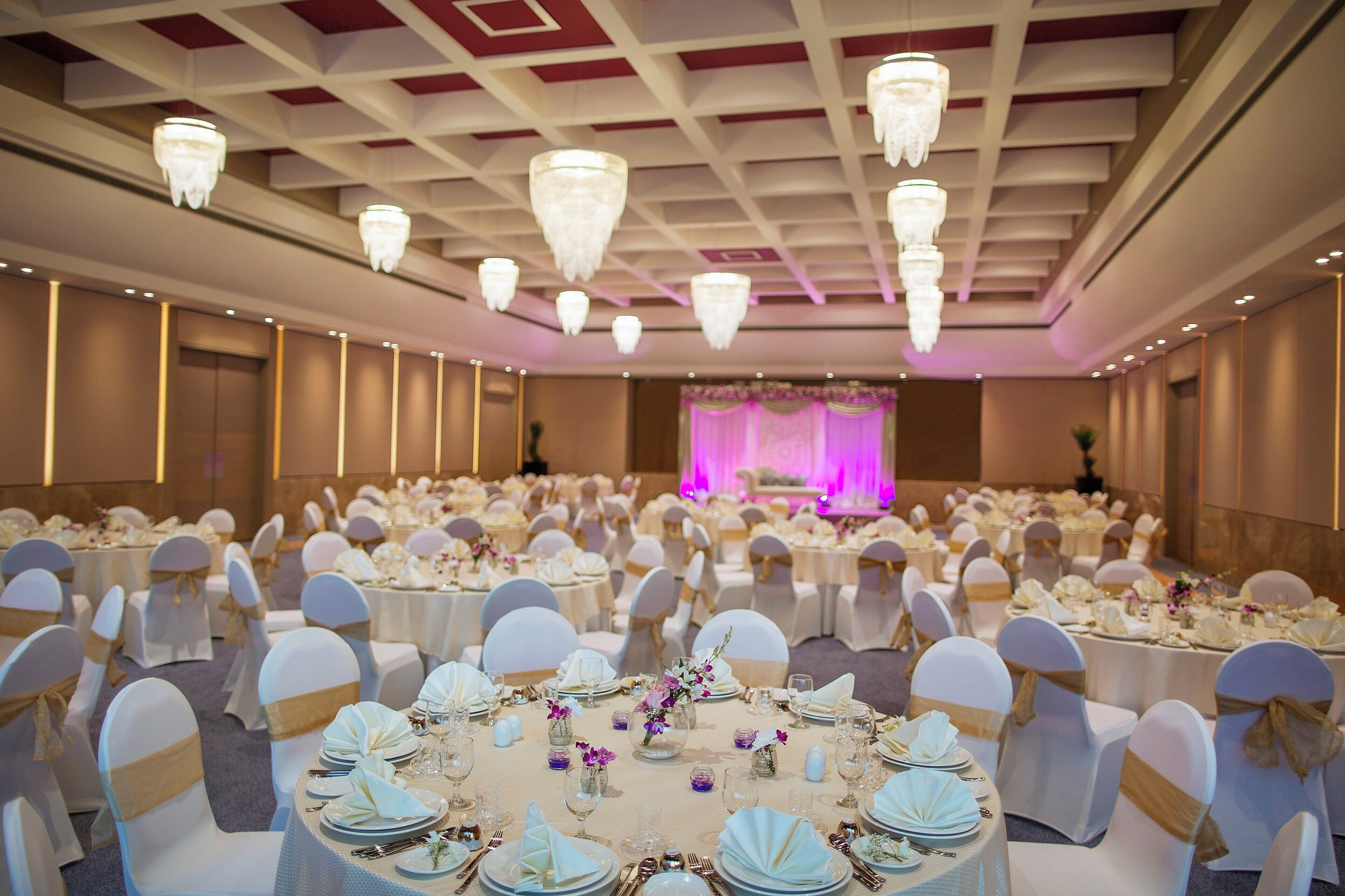 Ein Festsaal mit runden Tischen, festlich gedeckt für eine formelle Veranstaltung, mit weißen Tischdecken, goldenen Satinbändern und floralen Mittelstücken.