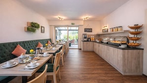 Un espace réservé au petit-déjeuner, avec une longue table, des sièges confortables, un comptoir où sont exposés les aliments, et une vue sur l’extérieur par la porte.