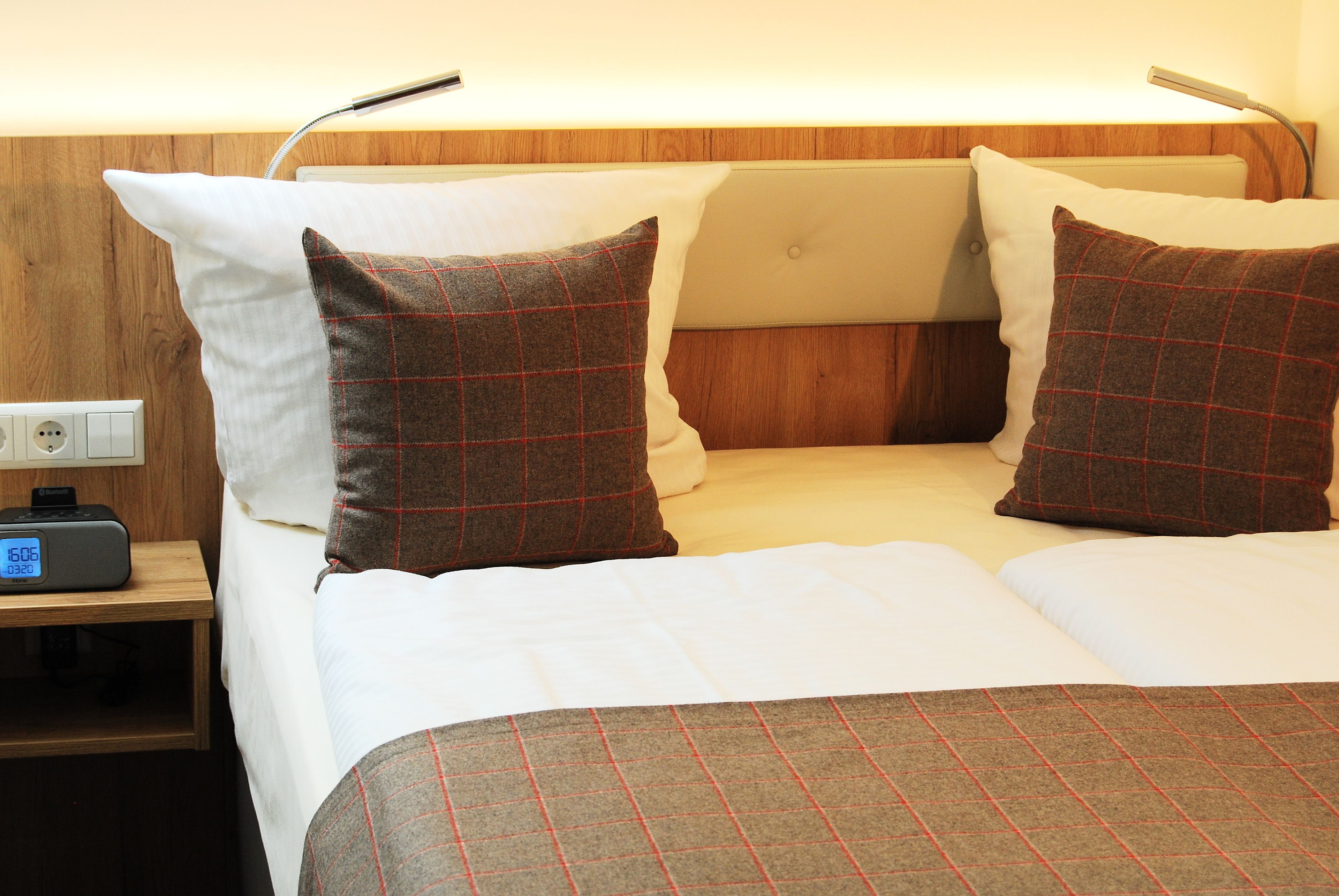 Uma cama bem arrumada com roupa de cama branca e xadrez, uma mesinha de cabeceira com abajur e uma tomada.
