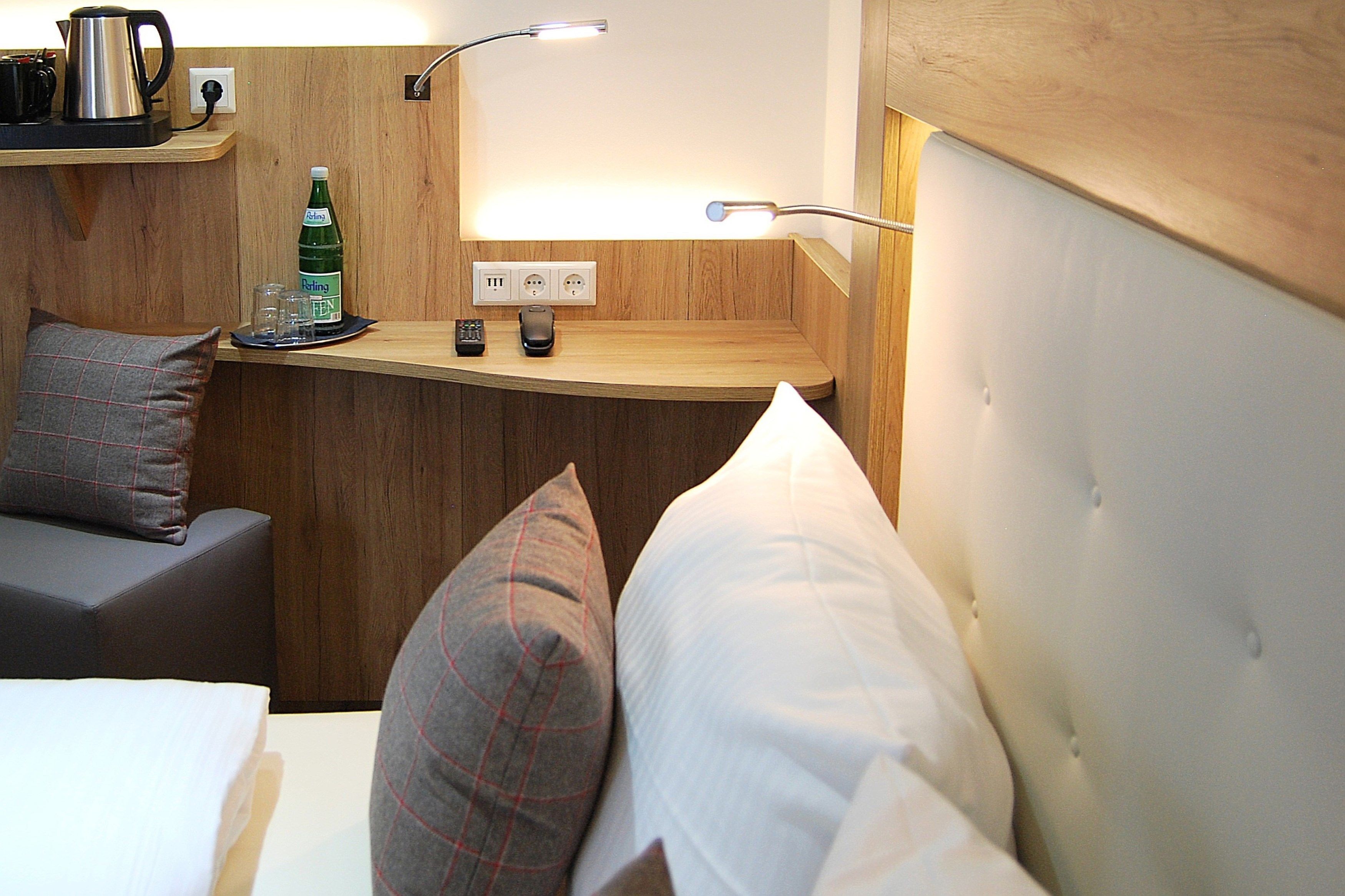 Una habitación de hotel compacta con cama, una mesita de noche con lámpara, un pequeño estante con un hervidor y una botella, y un enchufe.