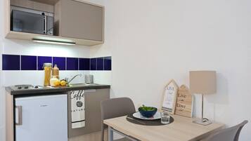 A compact kitchen with a microwave, sink, and a small refrigerator. There is a table with a bowl and a lamp.
