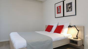 A neatly made bed with white and red pillows, a grey patterned blanket, and a bedside table with a lamp.