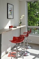 A modern room with a large window, a framed artwork on the wall, a potted plant, and two red bar stools.