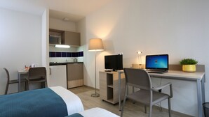 A hotel room with a bed, a desk with a computer, a chair, a lamp, a small kitchenette, and a potted plant. - Apparteo Nancy (Nancy)