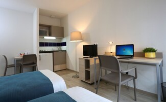 A hotel room with a bed, a desk with a computer, a chair, a lamp, a small kitchenette, and a potted plant.