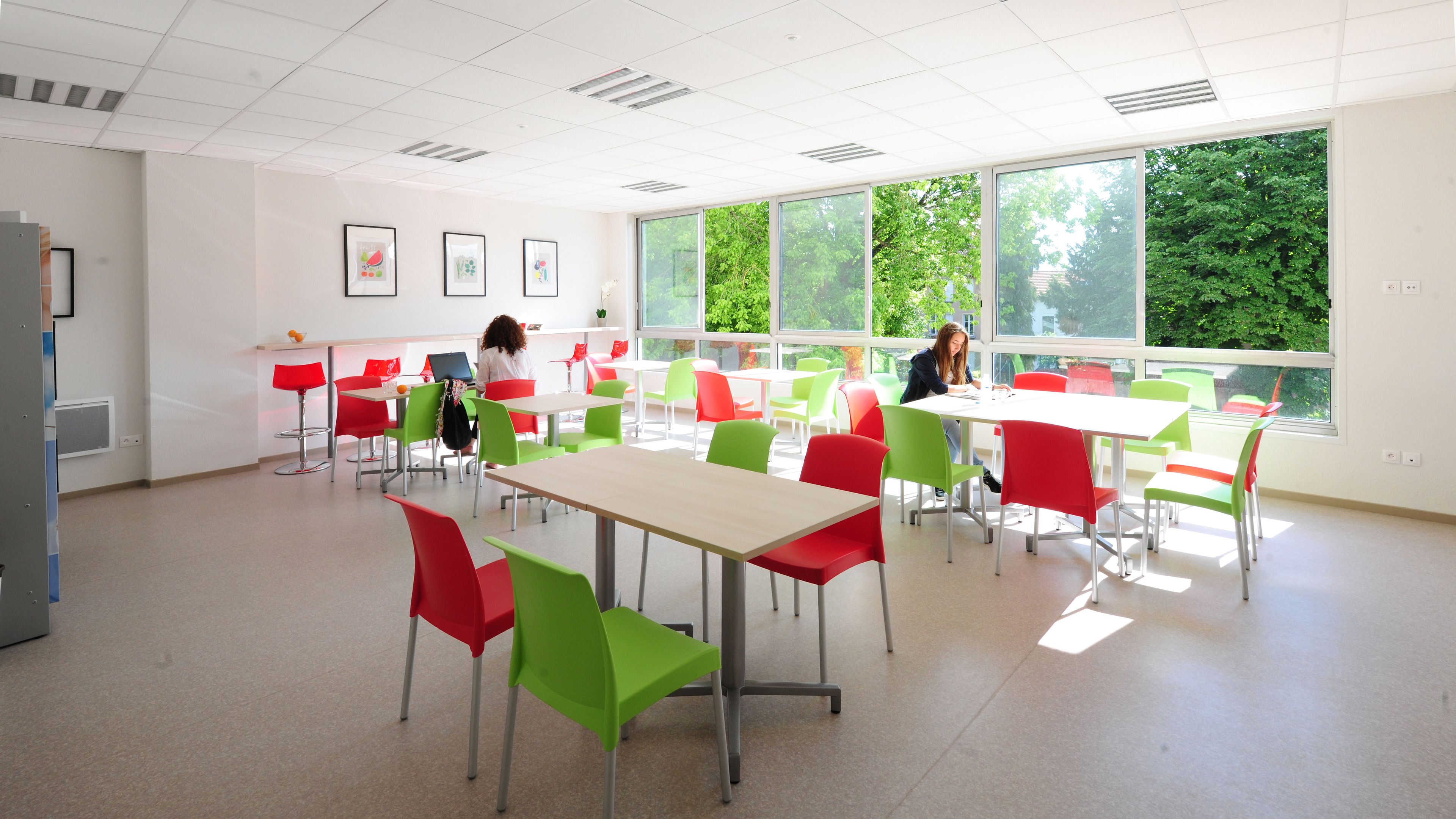 A classroom with tables and chairs, large windows, and artwork on the walls.