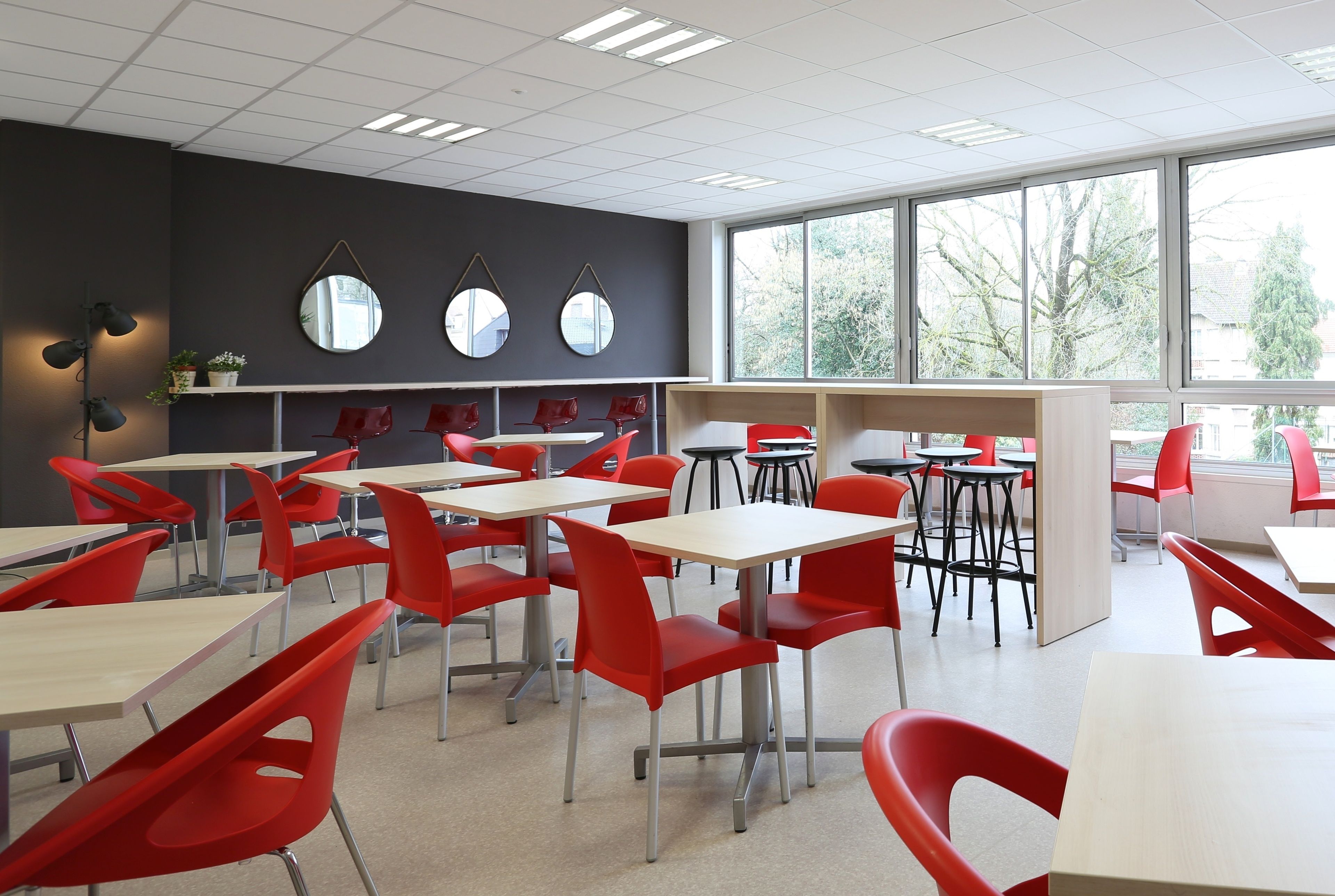 A classroom with red chairs and tables, large windows, and a dark wall with mirrors.