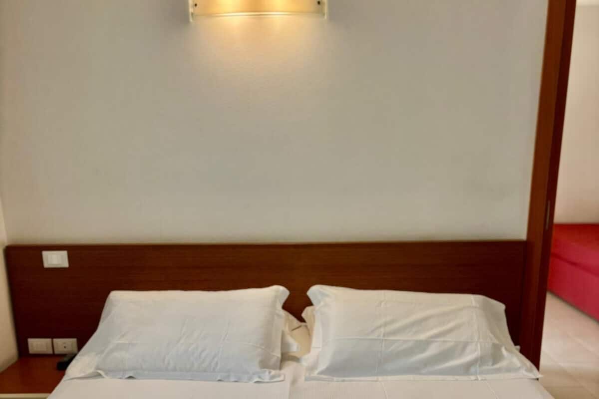 A neatly made bed with white linens, a wooden headboard, and a wall-mounted light fixture above it.